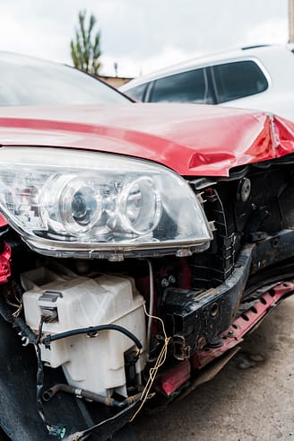 A selective focus shot of a damaged red car after a car accident, highlighting the crumpled fender and shattered headlight.