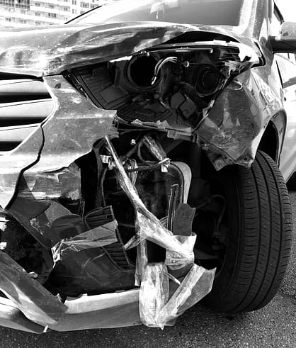 A disabled car with a crumpled hood and a flat tire sits on the side of a road after being involved in a collision.