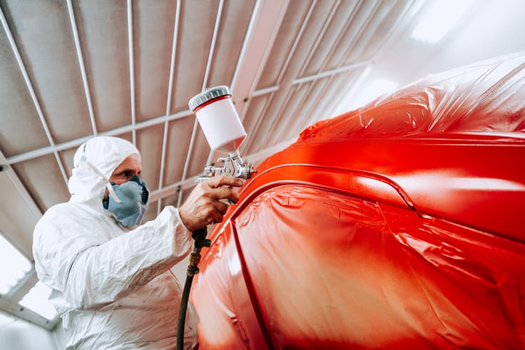 Automotive engineer wearing a protective suit and mask expertly spraying a red car with a paint gun in a paint booth.