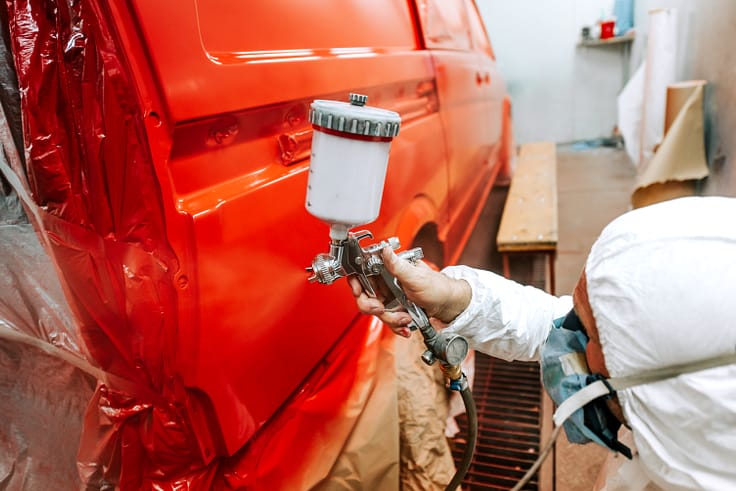 A professional painter in a protective mask and coveralls, expertly applying red paint to a car using a spray gun in a paint booth.