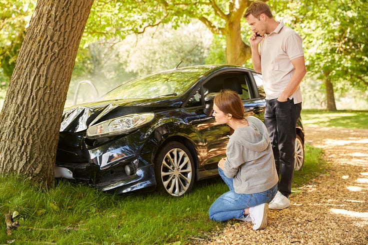 Two people stand by a wrecked car with a tree embedded in its front, one holds a phone to their ear while the other inspects the damage.