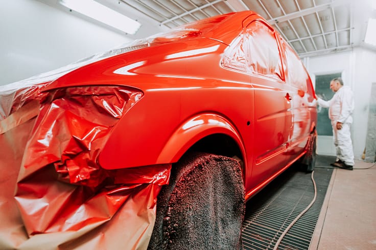 Worker wearing protective gear examining a car's new paint job under bright lights in a spray booth.