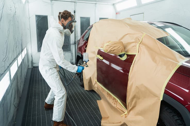 Worker in protective gear spraying degreaser on car door with spray gun.