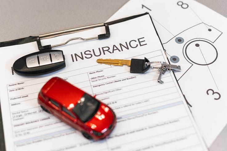 A hand holding a pen poised over a car insurance form, with a model car and insurance policy documents in the background.