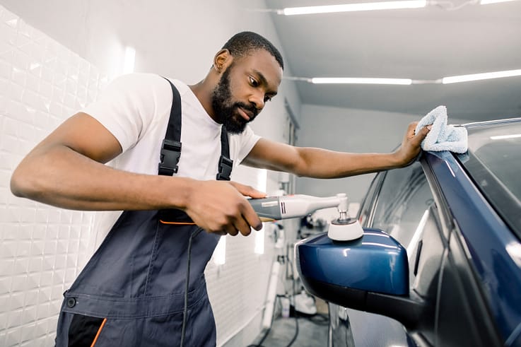 A focused auto detailer, wearing a white t-shirt and gray overalls, uses a polisher to restore the shine of a car's paintwork.