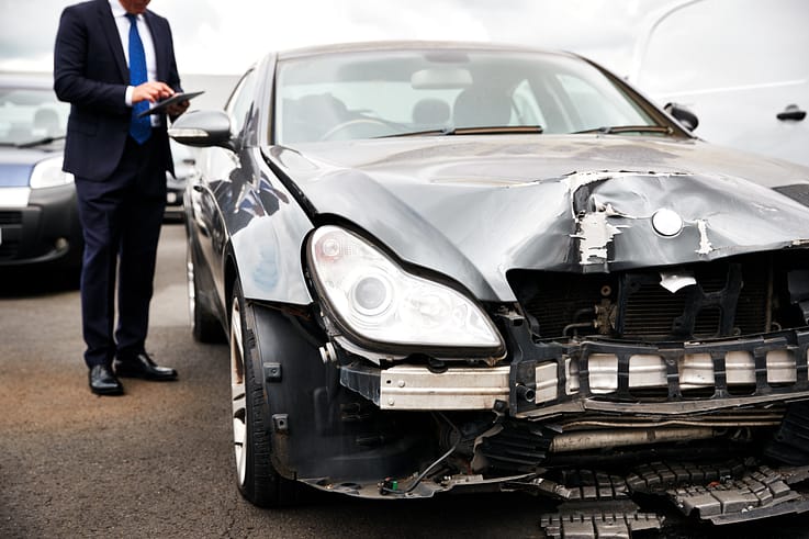 Insurance professional in a suit documenting vehicle damage with a tablet after a collision.