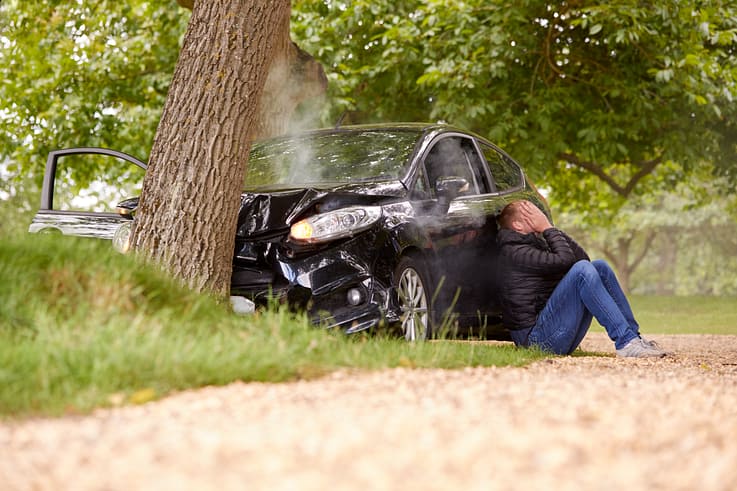 Distraught teenage driver sitting next to a damaged car after hitting a tree.