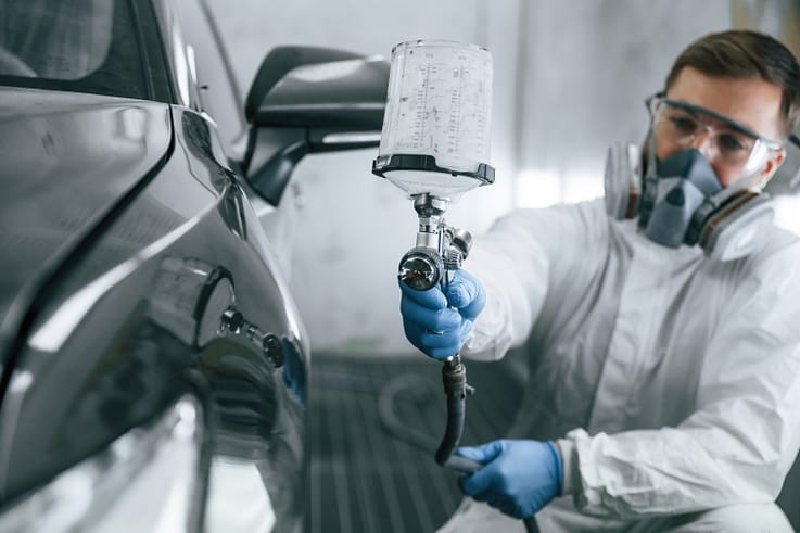 Worker in protective clothing carefully spraying paint onto a car in a well-ventilated paint booth.