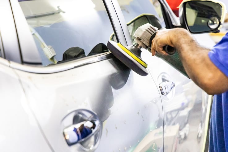 A worker is using a grinder machine to buff polish wax onto a car.