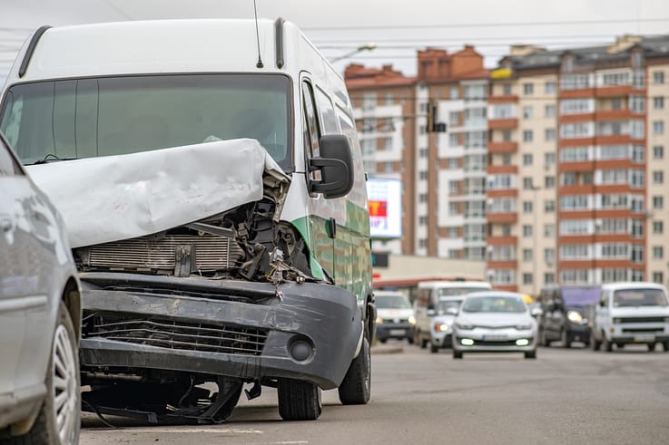 A heavily damaged car with a crumpled front end and shattered windshield sits abandoned on a city street after a collision.