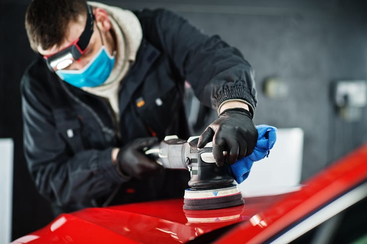 Car detailer in a face mask using an orbital polisher to buff out imperfections in a vehicle's paint.