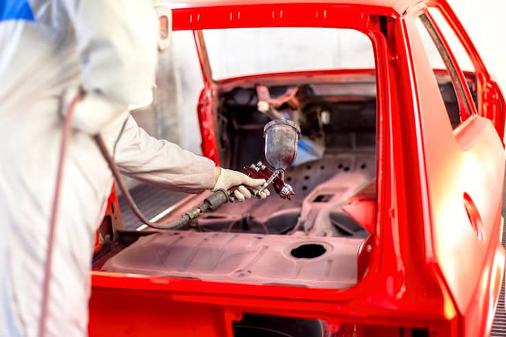 Skilled technician in a white protective suit applying red paint to a car in a professional paint booth.